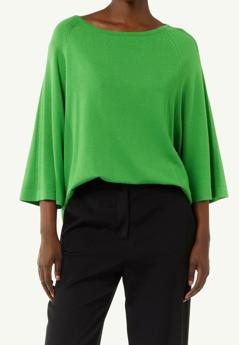 Femme portant un haut ample vert vif à manches larges et un pantalon noir taille haute sur un fond clair.
