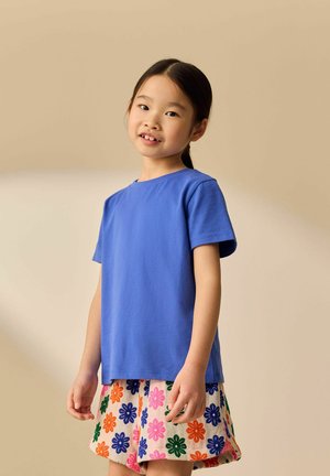 Jeune fille souriante, portant un t-shirt bleu et une jupe fleurie colorée, debout devant un fond beige uni.