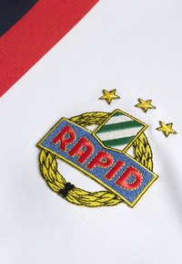 Hvidt stof med et struktureret mønster har et broderet logo, en gylden krans og tre stjerner over et grønt-stribet skjold, der er farvet i blå og rød.