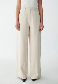 Beige wide-leg trousers with a smooth texture, featuring a high waist, belt loops, and discreet front pockets, paired with black pointed heels.