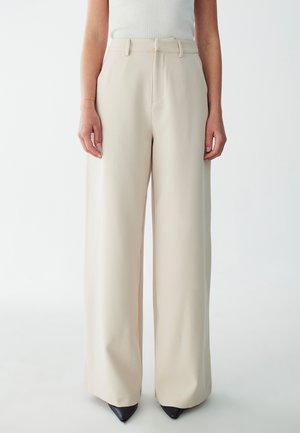 Beige wide-leg trousers with a smooth texture, featuring a high waist, belt loops, and discreet front pockets, paired with black pointed heels.