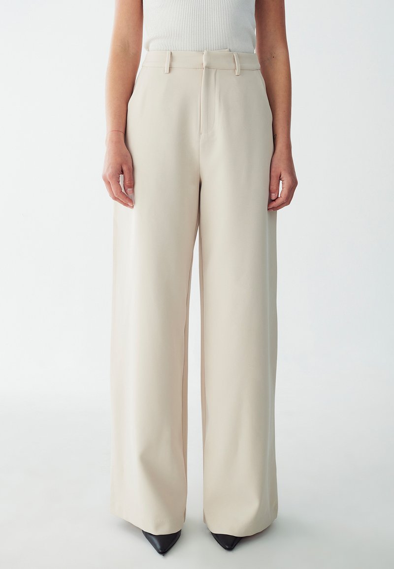 Beige wide-leg trousers with a smooth texture, featuring a high waist, belt loops, and discreet front pockets, paired with black pointed heels.