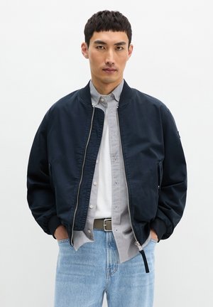 Jeune homme debout, les mains dans les poches, portant une veste bomber bleu marine, une chemise rayée, un t-shirt blanc, un jean bleu clair et une ceinture marron.