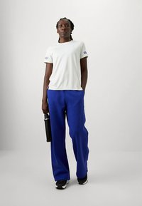 Valge T-särk sinise logodega, sellega sobitatud lahtised sinised püksid ja mustad tossud. Käes must veepudel.