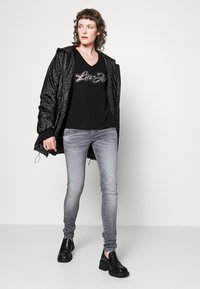 En svart pufferjakke med et subtilt mønster, lagdelt over en svart V-hals skjorte med glitrende bokstaver, sammen med lysegrå skinny jeans og svarte loafers.