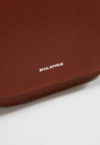 Bolso de cuero marrón con textura suave, que presenta el logotipo blanco "ROYAL REPUBLIQ" en la parte inferior derecha. Los bordes cosidos realzan el diseño.