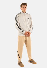 Chaqueta de entrenamiento blanca para hombre con franjas negras y logo, pantalones beige ajustados con bolsillos con cremallera y detalles negros, zapatillas marrones.