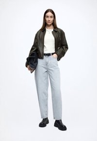 Jachetă bomber din piele verde închis, pulover alb, blugi cu talie înaltă, aspect de spălare deschisă, cizme negre masive, ținând o geantă neagră. Textură netedă.