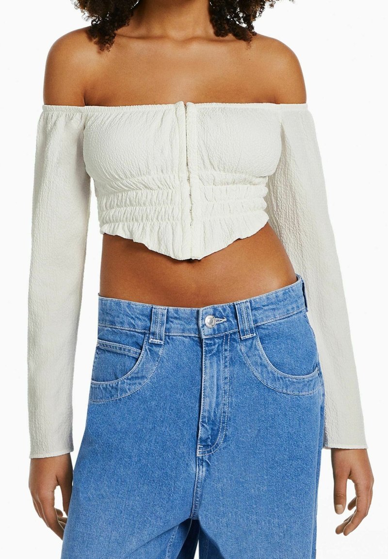 Donna che indossa un top cropped a maniche lunghe bianco con spalle scoperte e jeans in denim blu a vita alta e larghi, mostrata dalla gola ai fianchi.