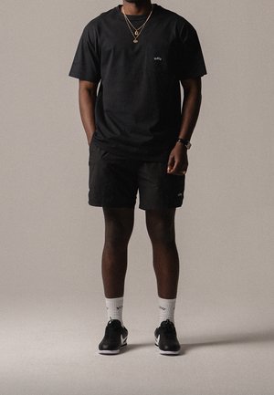 Schwarzes Baumwoll-T-Shirt mit Tasche, schwarze Shorts und schwarze Turnschuhe. Weiße Sneakersocken mit Logodetail. Einfaches und funktionales Design.