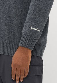 Kullfarget grå strikkegenser med ribbestrikkede mansjetter og nederkant. Har brodert tekst på ermet. Mykt materiale med en strukturert overflate.