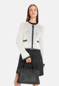White knitted cardigan with black accents and buttons, paired with a black leather handbag and a pinstriped skirt.