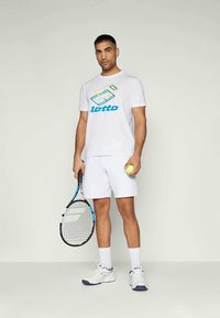 T-shirt atlético branco com o logotipo colorido da Lotto, combinado com calções brancos. O modelo segura uma raqueta e uma bola de ténis.