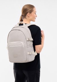 Sac à dos gris clair au toucher doux, avec des sangles réglables, une poche zippée à l'avant et un détail de logo. Une personne le porte par-dessus un t-shirt noir.