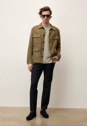 Mężczyzna w ciemnych okularach przeciwsłonecznych, oliwkowozielonej kurtce z przednimi kieszeniami, jasnoszarej koszuli, czarnych spodniach i czarnych butach, stojący na tle jednolitej ściany.