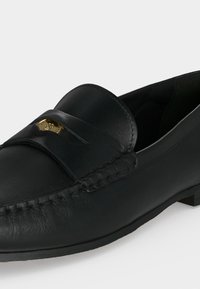 Μαύρο δερμάτινο loafer με ραφές μοκασίνου και χρυσό διακοσμητικό έμβλημα στο λουράκι, με χαμηλό συσσωρευμένο τακούνι και λεία σόλα.