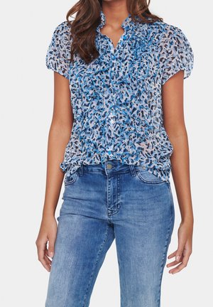 Kobieta nosząca niebieską i czarną bluzkę w zwierzęcy wzór z falbankami na krótkich rękawach, w połączeniu ze średnio wypranymi niebieskimi dżinsami na białym tle.