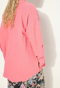 Pink lightweight shirt with a textured finish, featuring a collar and long sleeves, paired with black floral patterned pants.
