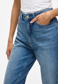 Jean en denim bleu taille haute avec une jambe droite, comportant une poche avant, des coutures discrètes et une fermeture à bouton en métal.