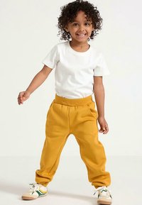 Pantaloni della tuta gialli con elastico in vita, t-shirt a maniche corte bianca e sneakers bianche con dettagli verdi e beige. Bambino in posa giocosa.