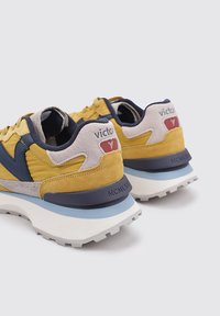 Sneaker gialli e grigi con dettagli blu navy; materiali in suede e mesh; suola bianca chunky; logo sul tallone; dettagli superficiali texture.