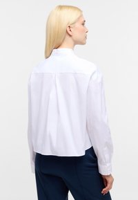 Hvid skjorte med lange ærmer, detaljer med rygfold og knaplukning ved ærmerne. Glat bomuldsstof, kortet design, minimalistisk stil.