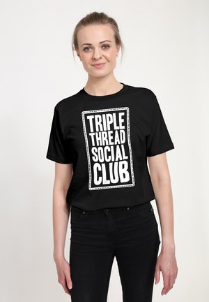 Kvinde med lys hud og hår trukket tilbage iført en sort t-shirt med hvid tekst "TRIPLE THREAD SOCIAL CLUB" står foran en ensartet, lys baggrund.
