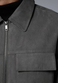 Chaqueta de ante gris con cuello, cierre frontal y un bolsillo de parche. Textura suave y diseño minimalista.