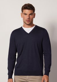 Marineblauer V-Ausschnitt-Pullover mit feiner Stricktextur, langärmlig und mit gerippten Bündchen. Getragen über einem weißen Rundhals-T-Shirt.