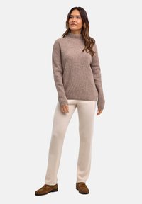 Brązowy prążkowany sweter z wysokim dekoltem, zestawiony z kremowymi spodniami i brązowym obuwiem. Sweter ma długie rękawy i luźny krój.