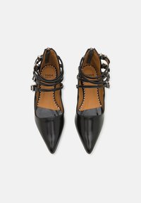 Talons en cuir verni noir avec des pointes pointues, de multiples brides croisées et des boucles en argent. L'intérieur présente une semelle intérieure marron.