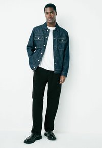 Denim jacket in dark blue with two front pockets, silver buttons, worn over a white shirt, paired with black pants and black shoes.