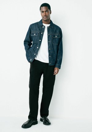 Dunkelblaue Jeansjacke mit zwei vorderen Taschen, silbernen Knöpfen, getragen über einem weißen Hemd, kombiniert mit schwarzen Hosen und schwarzen Schuhen.