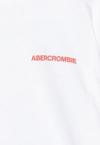 Bílý bavlněný tričko s logem "ABERCROMBIE" v korálově červené barvě. Design je jednoduchý, s kulatým výstřihem a krátkými rukávy.