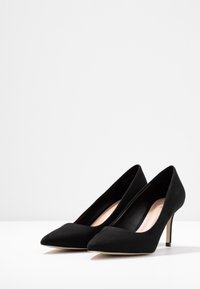 Escarpins en daim noir à talons hauts avec pointe effilée, talon stiletto fin, et doublure intérieure lisse. La semelle est de couleur plus claire, contrastant avec la tige.