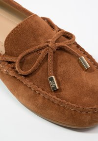 Mocassin en daim marron avec coutures apparentes, nœud noué sur le dessus et embouts métalliques dorés gravés d'un logo.