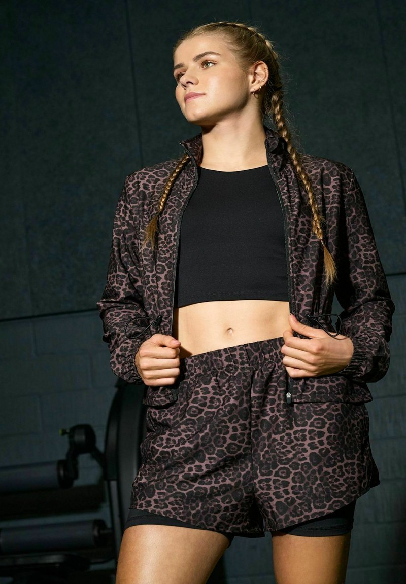 Femme aux cheveux tressés portant un crop top noir, une veste à motif léopard et un short assorti sur un fond sombre de salle de sport.
