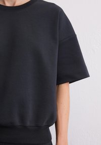 Donkergrijs sweatshirt met korte mouwen, ronde halslijn en geribbelde zoom. Gladde textuur en minimale stikseldetails op de mouwranden.