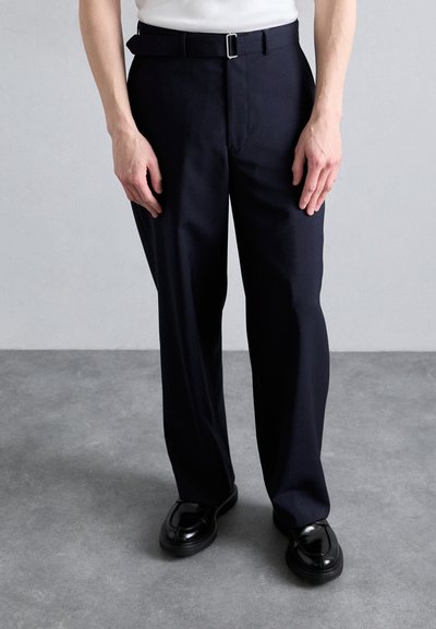 Pantaloni negri cu un design drept, prevăzuți cu o curea cu o cataramă argintie. Țesătură netedă și detalii minime. Pantofi negri dedesubt.