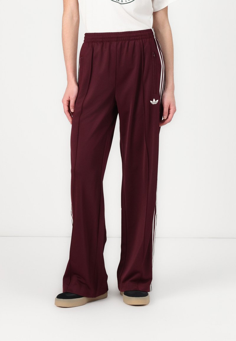 Pantaloni sportivi de culoare burgund cu o croială relaxată, dungi laterale albe, buzunare cu fermoar și un logo brodat pe șoldul stâng.