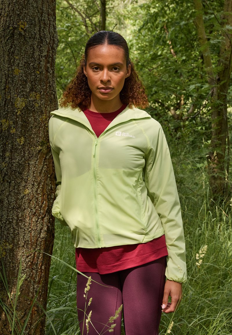 Giacca zip-up verde chiaro con vestibilità rilassata, indossata sopra una maglietta rossa e leggings viola, con uno sfondo naturale verde.