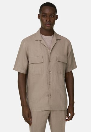 Mies pukeutuneena beigeen, lyhythihaiseen nappipaitaan, jossa on kaksi etutaskun läppää, ja vastaavat housut yksivärisellä taustalla.