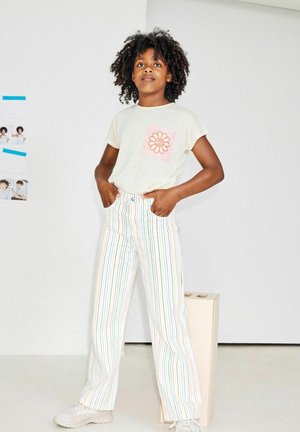 T-shirt crème avec une poche en forme de fleur rose, associé à un pantalon large à rayures multicolores. Le modèle se tient les mains sur les hanches, portant des baskets claires.
