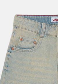 Jean en denim bleu clair avec des boutons en cuivre, présentant une petite poche avant et un léger dégradé. Étiquette de la marque visible à l'intérieur de la ceinture.