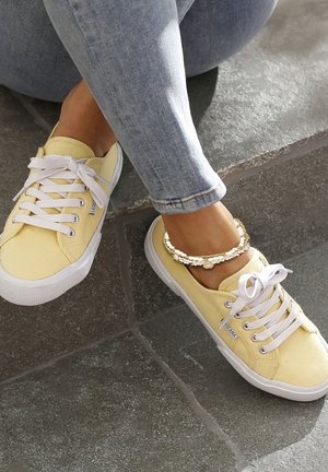 Yellow canvas sneakers with white laces and rubber soles, featuring a logo patch on the side. Paired with light blue fitted jeans.
