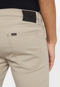 Närbild av beige Lee-jeans bakfickan med varumärkesetikett och en persons högra arm som vilar vid höften.