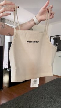 Beige Cropped-Top mit dünnen Trägern, gehalten von tätowierten Armen, die den Text "PEGADOR" zeigen und einem beigefügten weißen Etikett in einem Wohnzimmer.