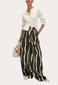 Femme portant un cardigan blanc côtelé à boutons, un pantalon noir à jambes larges avec des rayures verticales blanches, des chaussures blanches tissées et portant un sac à main beige.