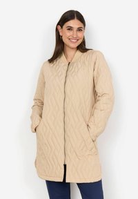 Beige quiltad jacka med dragkedja fram, ribbat krage och sidofickor. Har en lång, rak passform och diamantmönstrad textur.