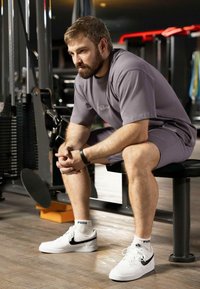 Maglietta atletica grigia e pantaloni corti, seduto su una panchina in una palestra. Indossa scarpe Nike bianche con accenti neri e calze Puma. Texture del pavimento in legno.